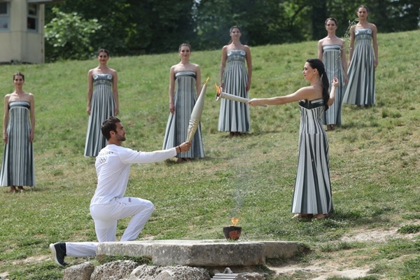 扮演最高女祭司的演員瑪麗·米娜（前右）點(diǎn)燃第一棒火炬手——希臘運(yùn)動(dòng)員、東京奧運(yùn)會男子賽艇項(xiàng)目金牌得主斯特凡諾斯·恩圖斯科斯手中的火炬。新華社記者 趙丁喆 攝